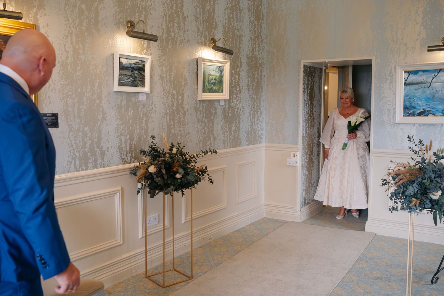 Groom in blue suit watching bride enter elegant wedding venue hallway.