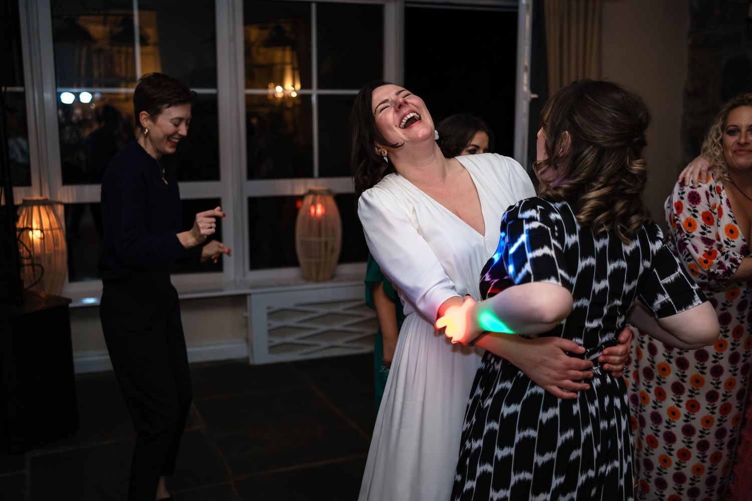 Bride in white dress laughing joyfully while hugging a guest on the wedding reception dance floor.