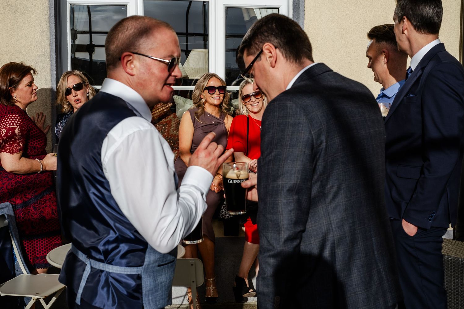 Wedding guests mingle outdoors in sunny weather, with a man in a vest checking his phone.
