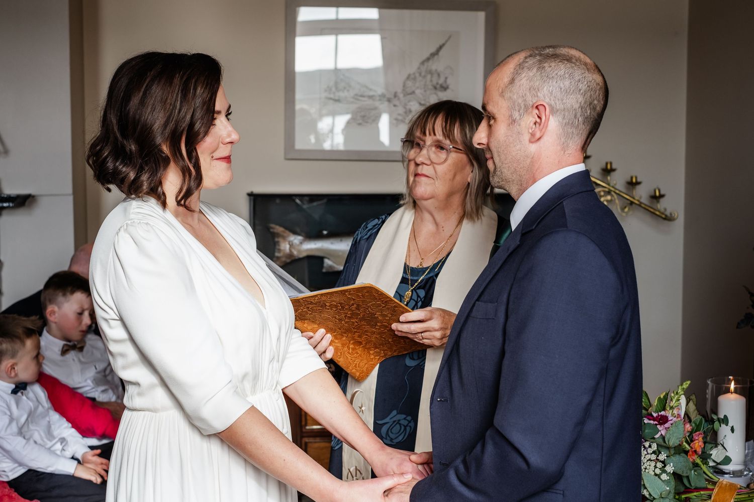 Bride and groom exchange vows indoors, officiated by a woman holding a brown book during an intimate wedding ceremony.