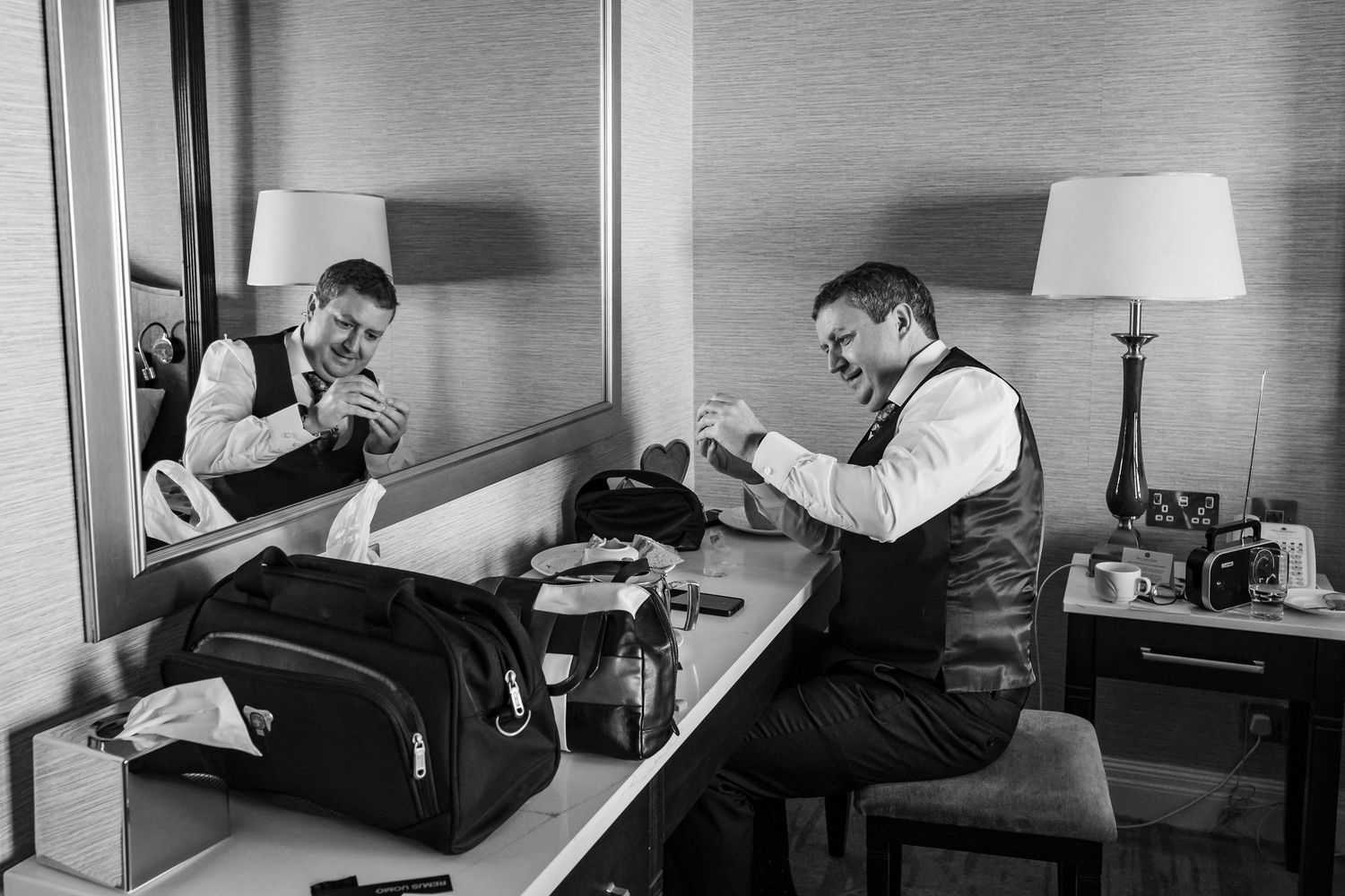 Groom getting ready in hotel room, adjusting cufflinks at mirror-side desk with travel bag in black and white photo.