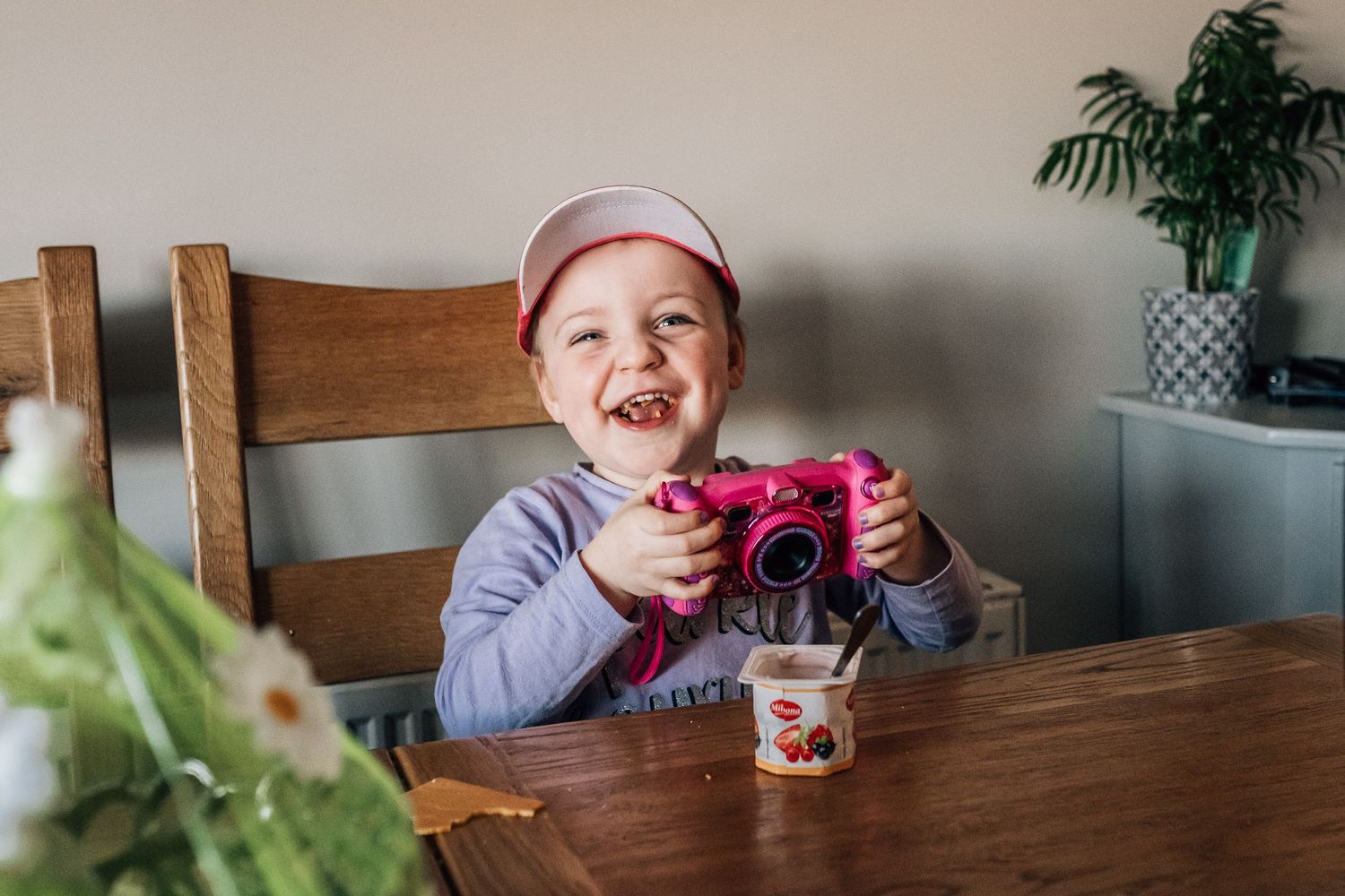 Child sitting at wooden table holds pink camera with houseplant in background.