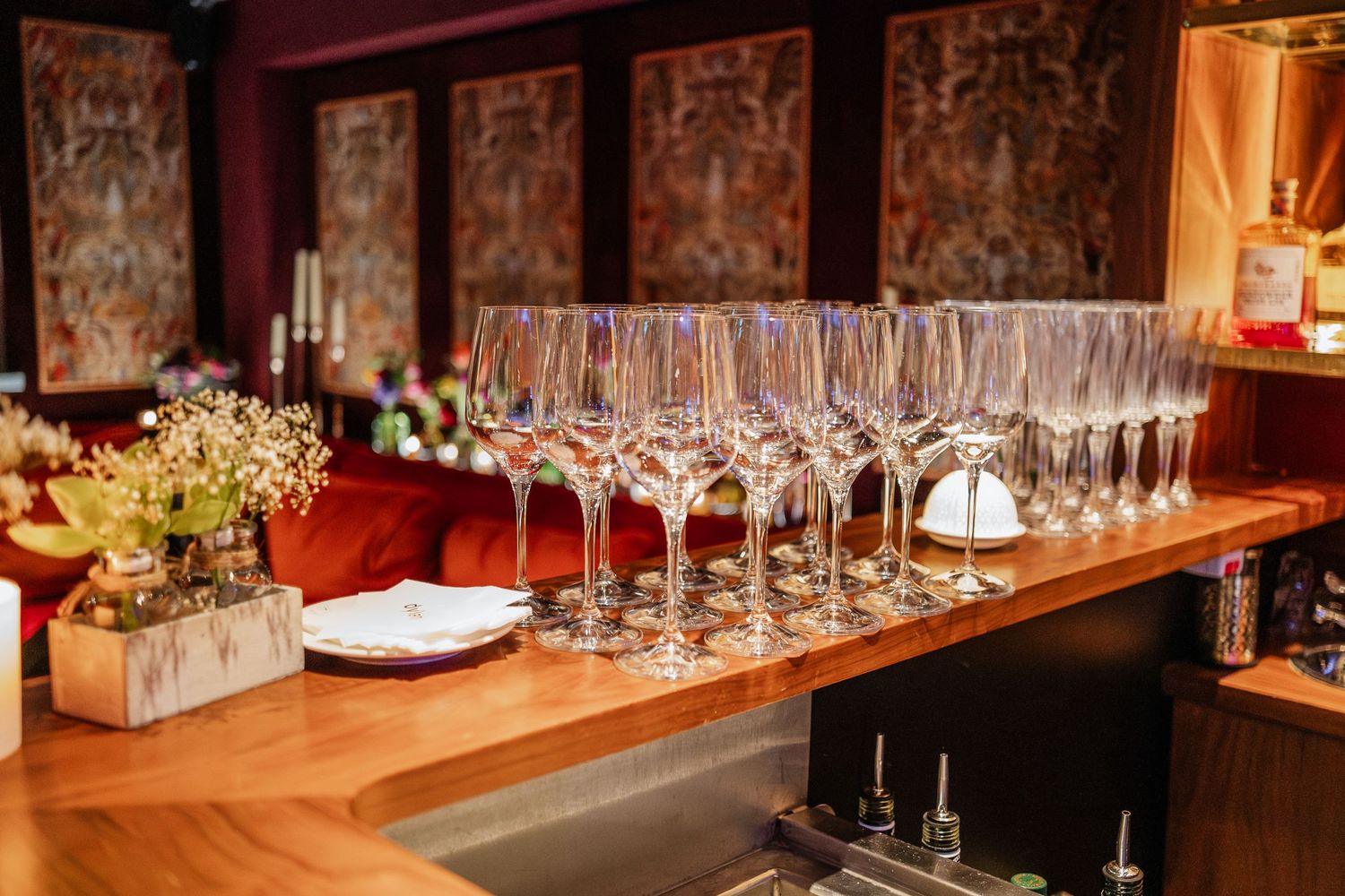Empty wine glasses lined up on a wooden bar counter with floral decorations.