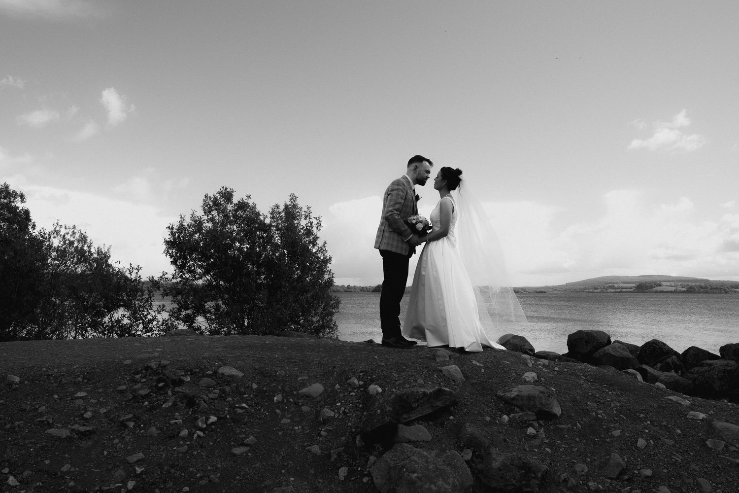 Black and white photo of a bride and groom sharing a romantic moment by the ocean with mountains in the background.