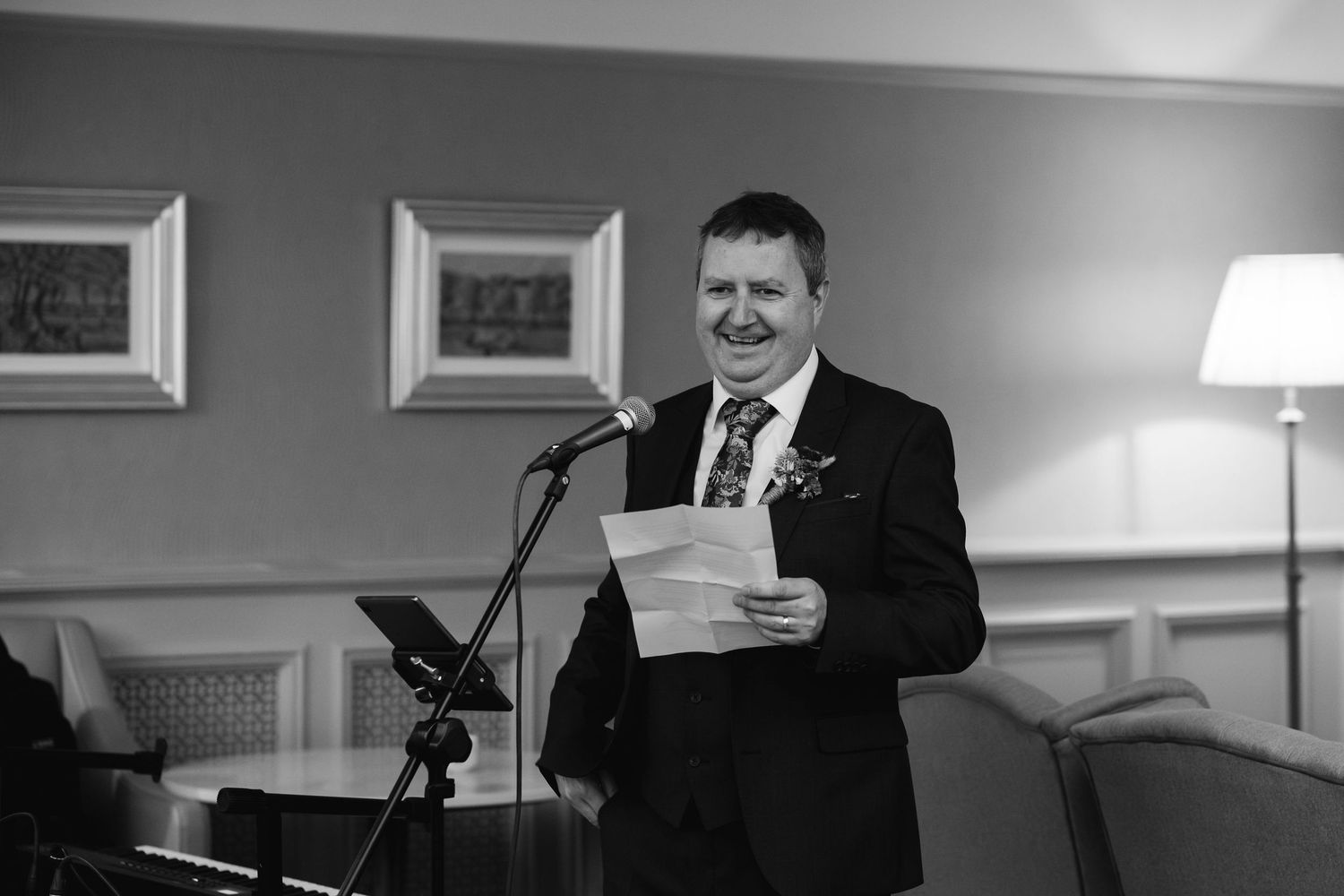 Man in suit smiling at microphone while holding papers, delivering speech at formal event, black and white photo.