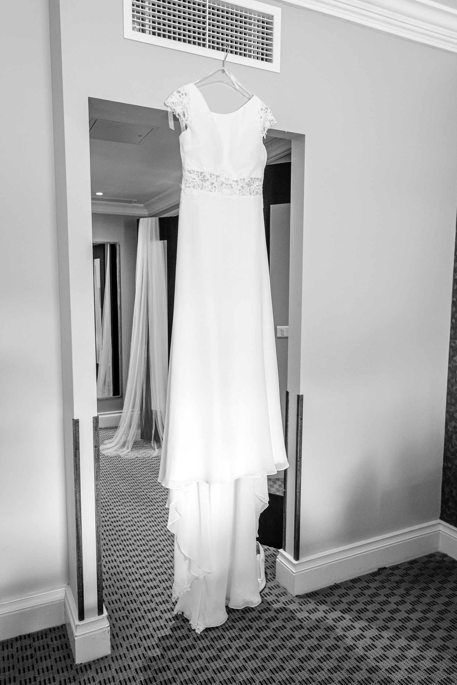 White wedding dress hanging in a hotel room with patterned carpet and white walls.