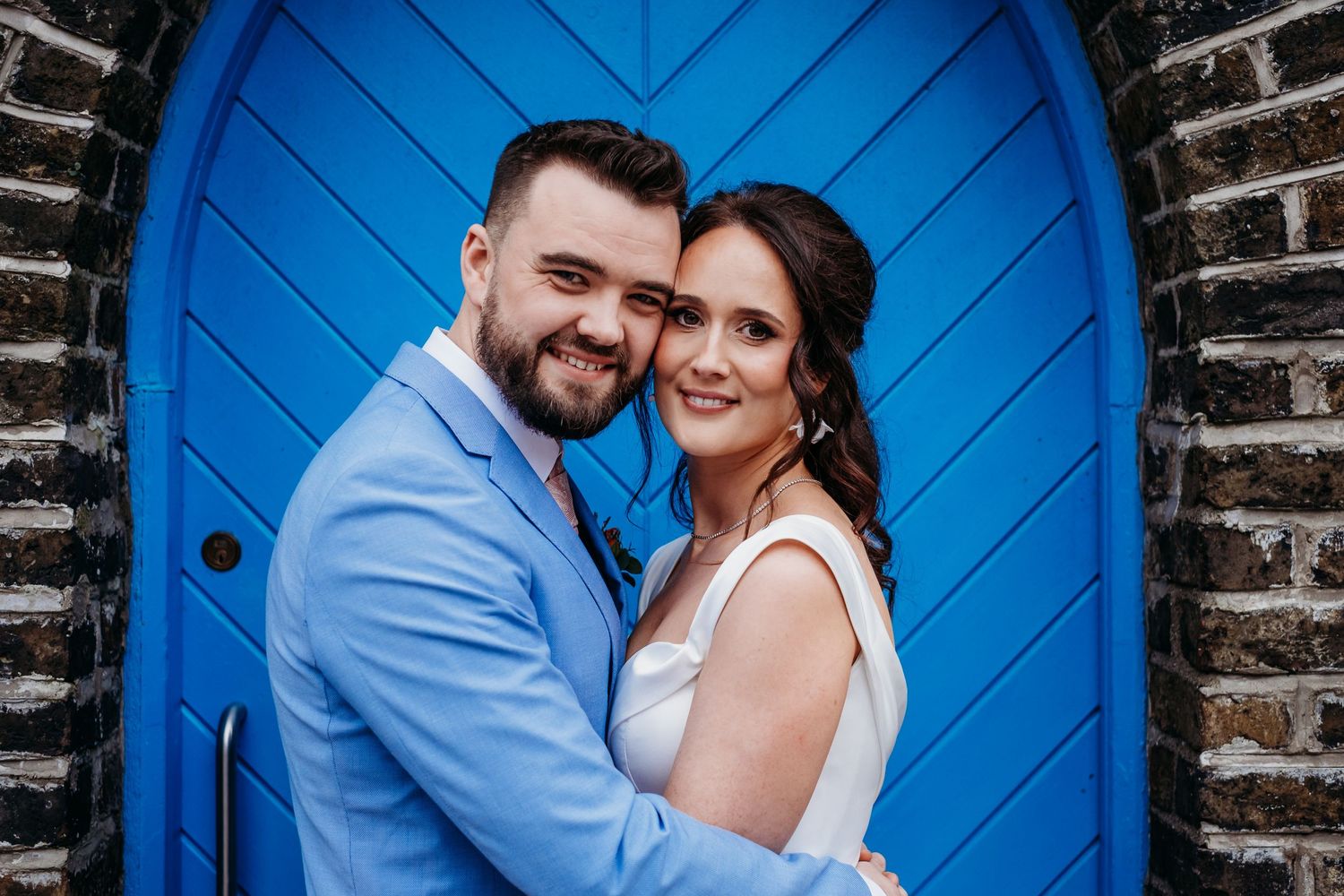 A romantic couple poses in front of a bright blue door against a brick wall on their wedding day.