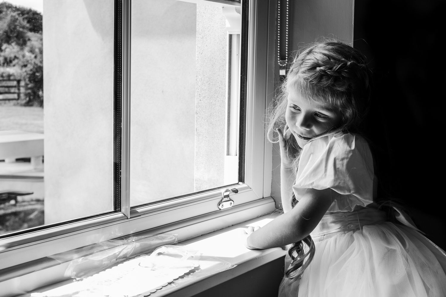 Black and white photo of a young flower girl in white dress looking out of window.