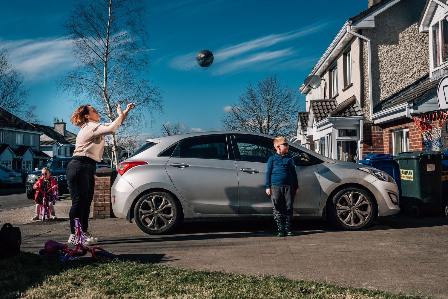 People playing catch with a ball in front of a silver car on a sunny day.