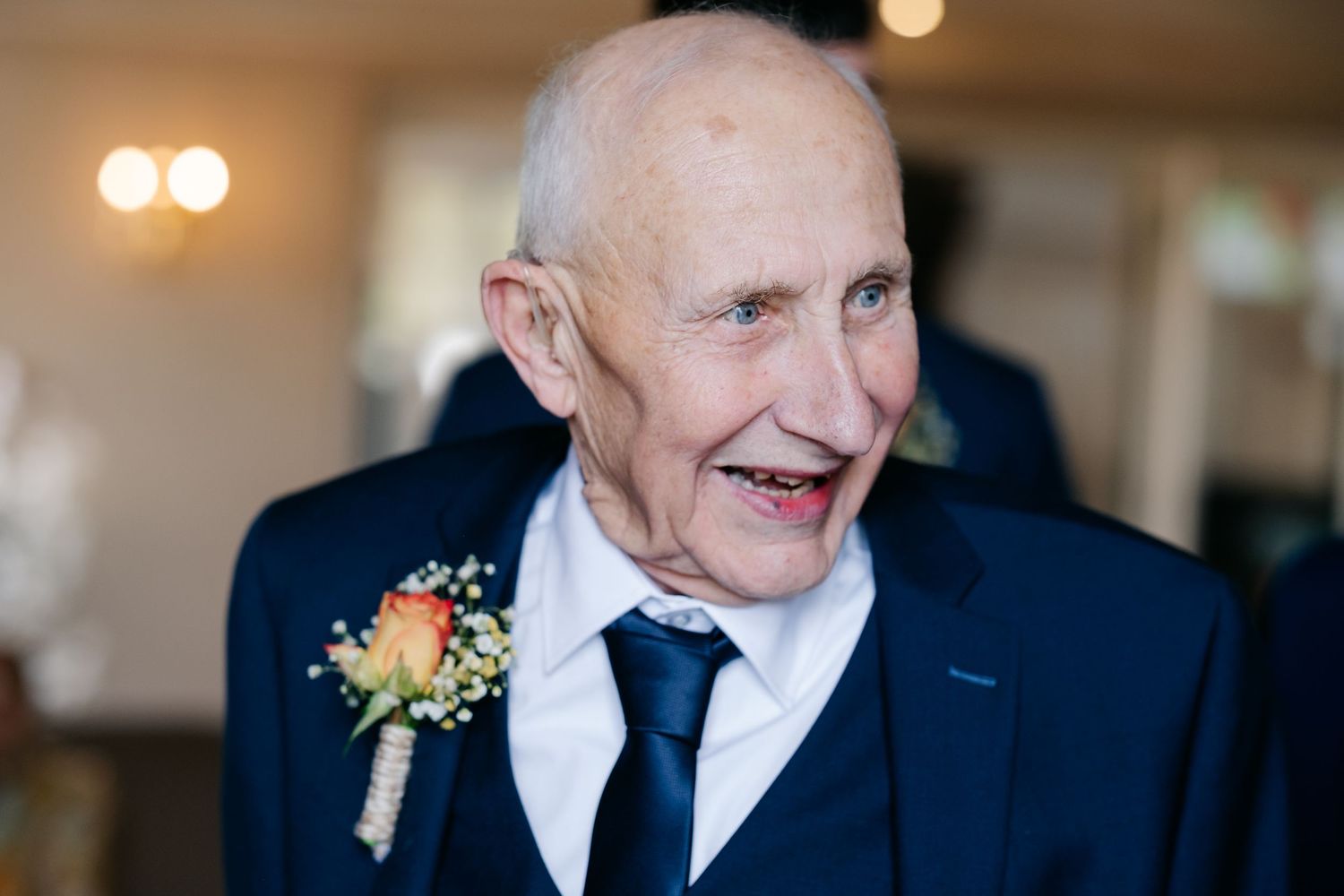 A person in a dark suit smiles warmly while wearing a boutonniere at a formal event.