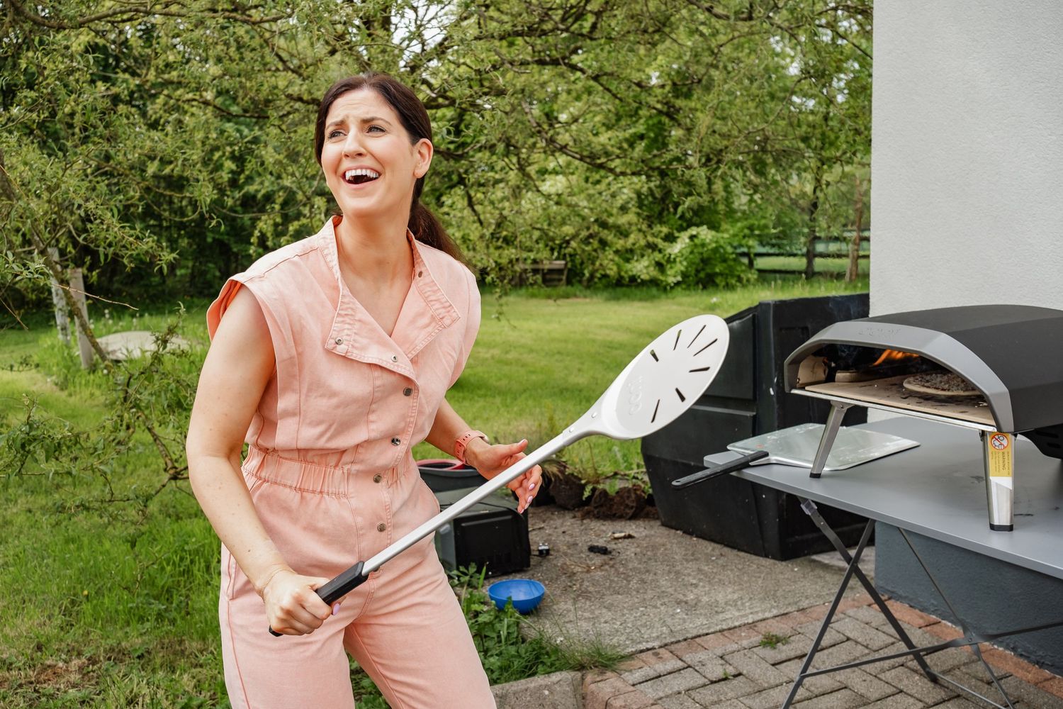 Person in pink outfit holds cooking utensil while standing near outdoor grill.