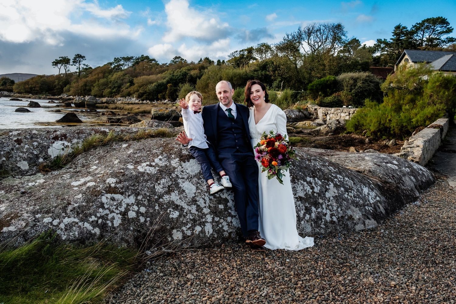Bride and groom pose with child on rocky coastal shoreline, bride holding colorful bouquet on overcast day.