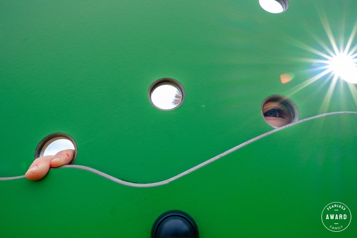 Close up of a child with peeping eye and fingers through a green structure at a playground.