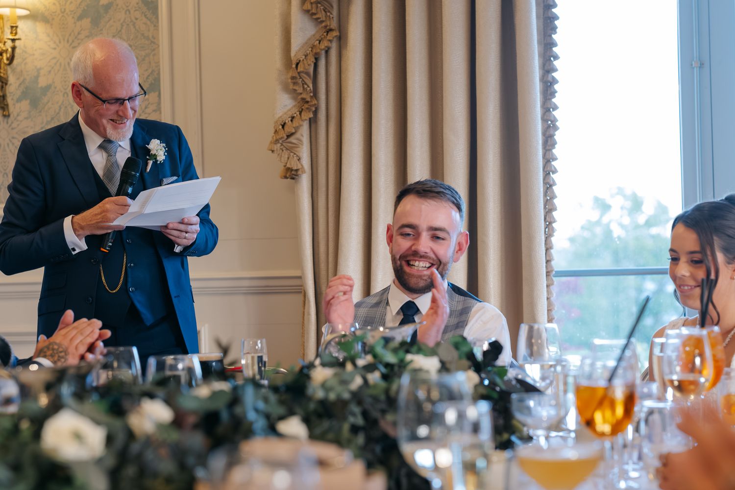 Groom laughing during wedding speech at elegant reception, man in blue suit reading from paper at head table.