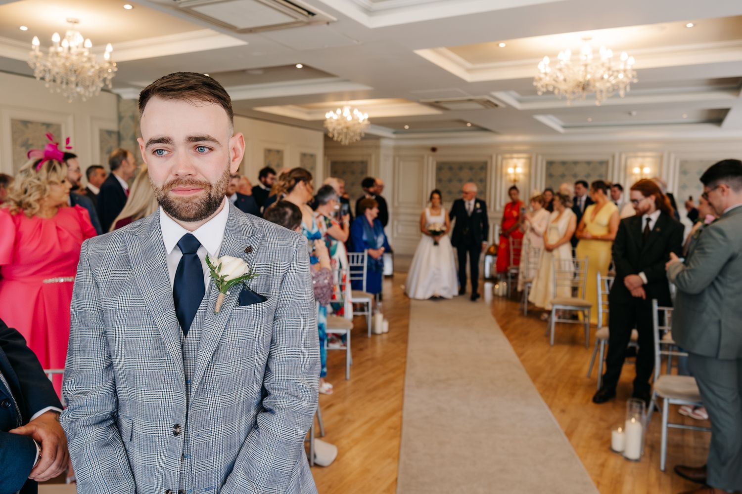 Groom in gray checked suit stands at altar as bride walks down aisle in elegant wedding venue.