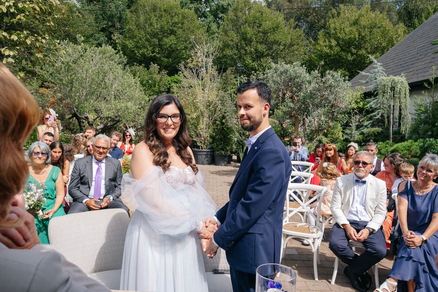 Bride and groom holding hands at outdoor wedding ceremony with guests seated in white chairs among lush greenery.