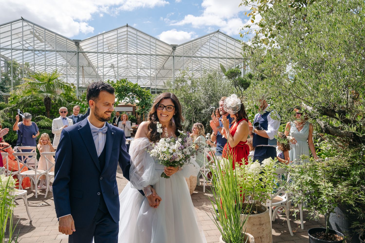 Bride and groom walk down outdoor aisle at greenhouse wedding venue surrounded by guests and lush greenery.