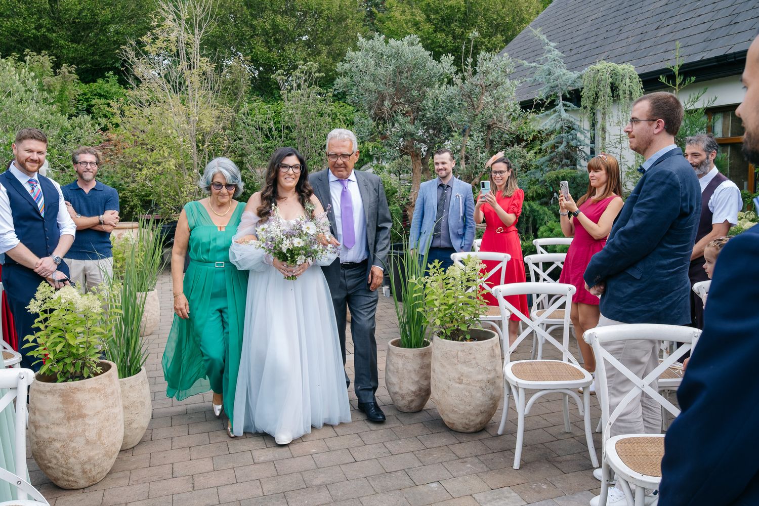 Bride walks down outdoor aisle with family at garden wedding ceremony, guests applauding in background.
