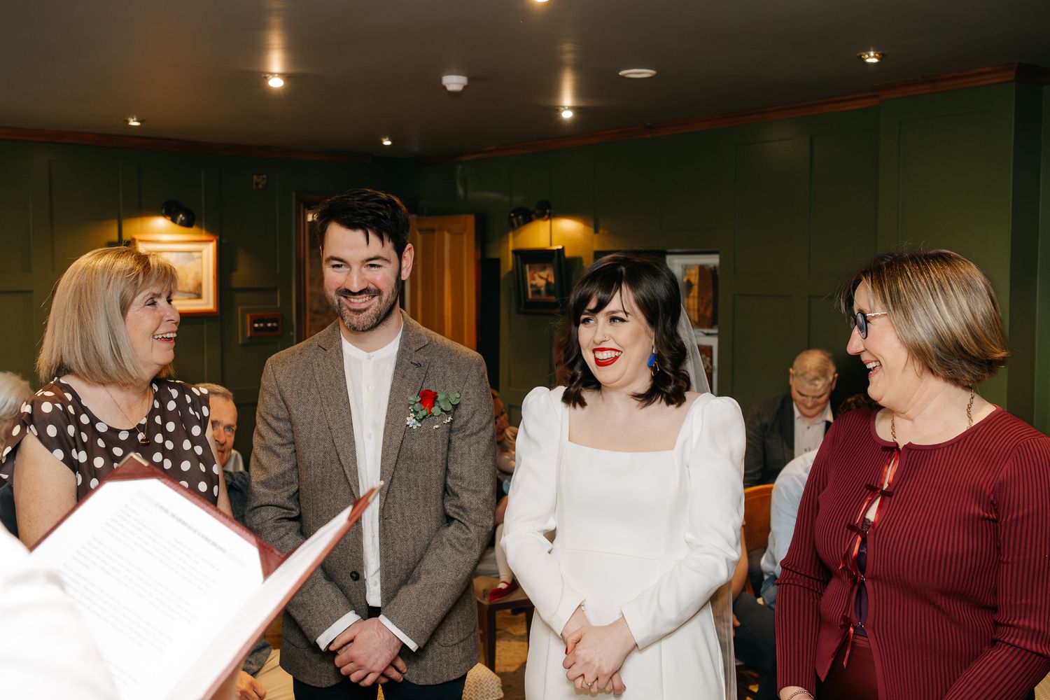 Bride in white dress and groom in tweed suit smiling with guests during indoor wedding ceremony.
