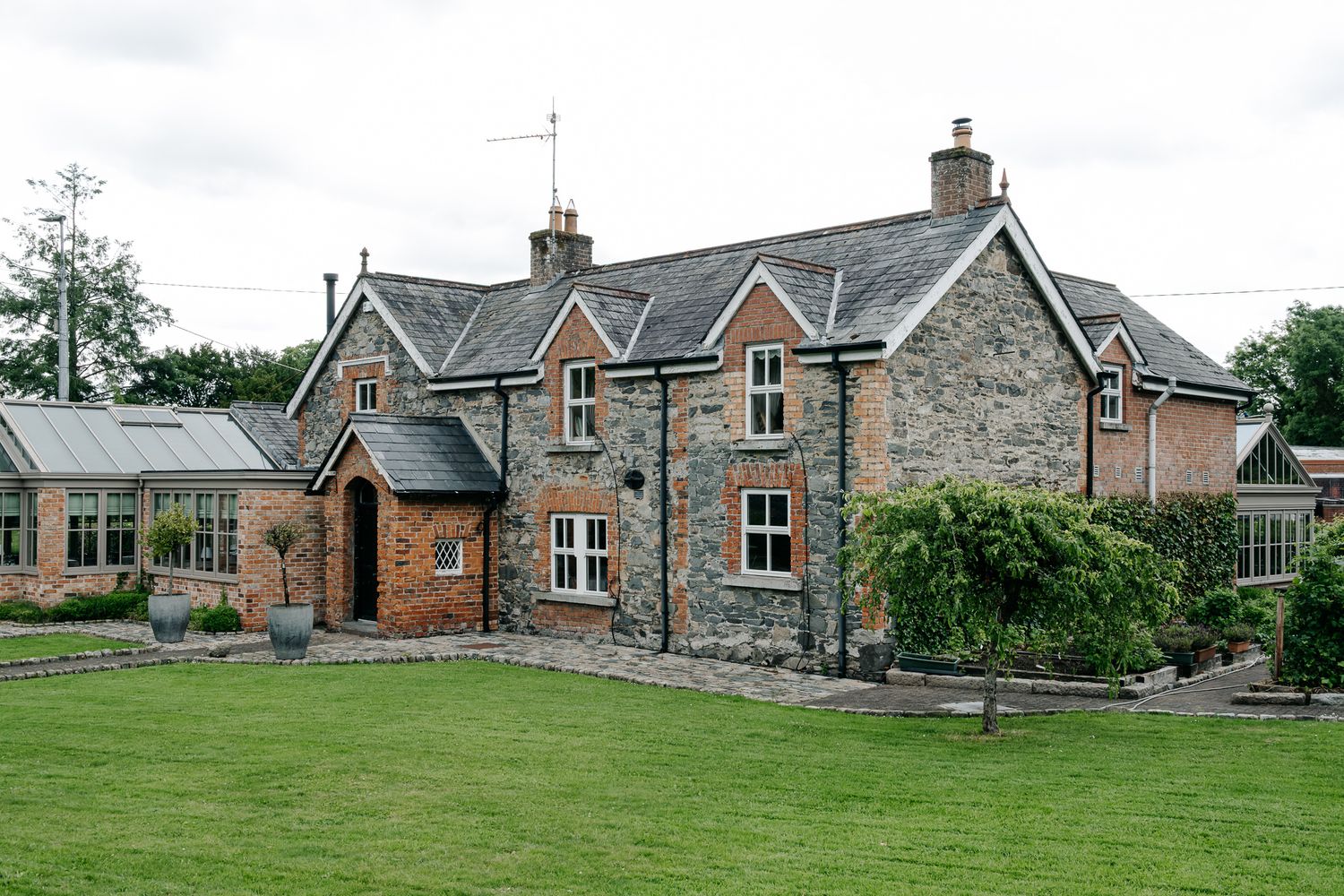 Charming stone and brick countryside cottage with slate roof, lush green lawn, and mature trees in a rural English setting.