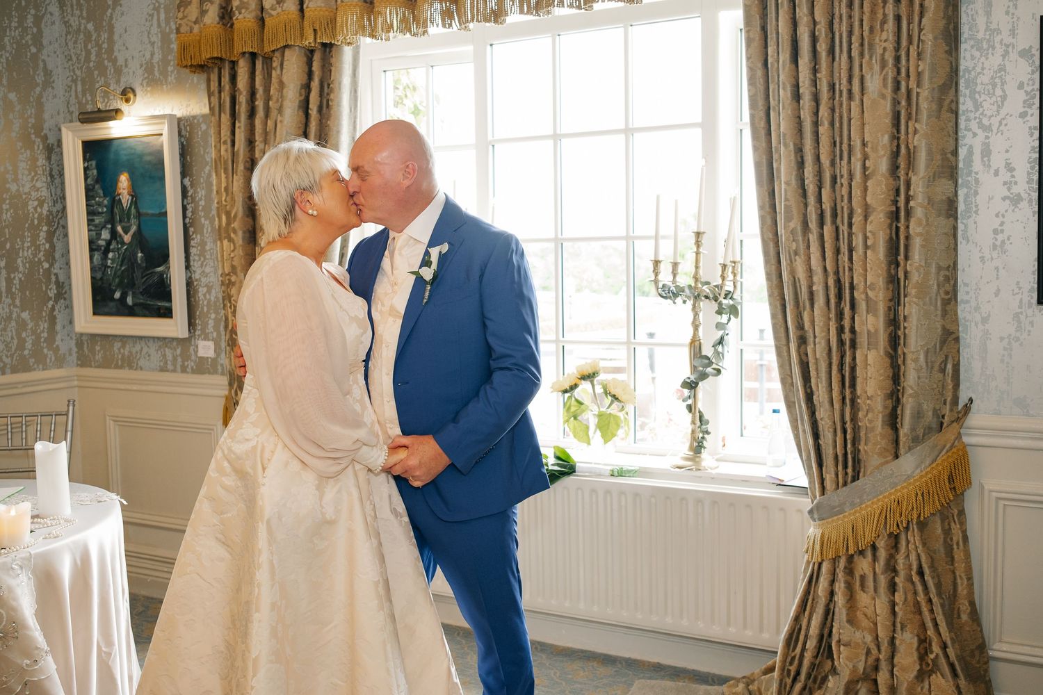Bride and groom share their first kiss by a window in an elegant room after their wedding ceremony.