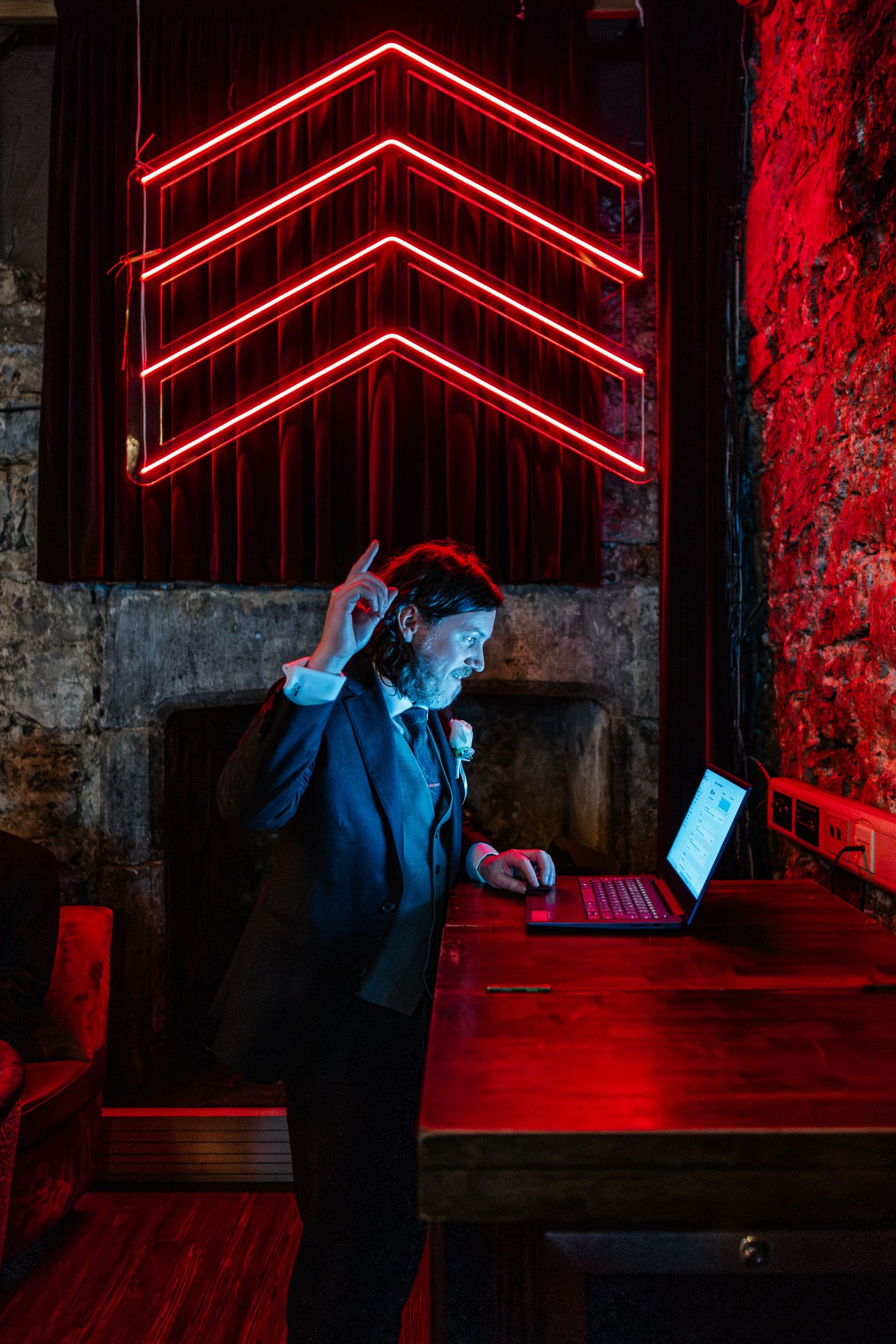 Man in suit using red laptop under neon chevron signs in moody red-lit bar environment.