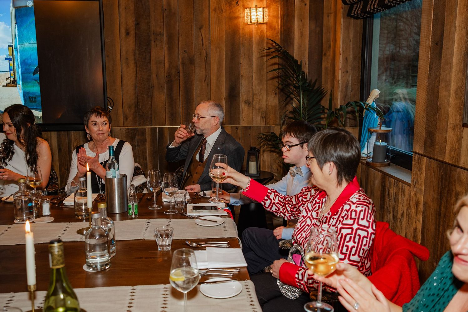 Group of people raising glasses for a toast at a festive dinner party in a warmly lit wood-paneled restaurant.