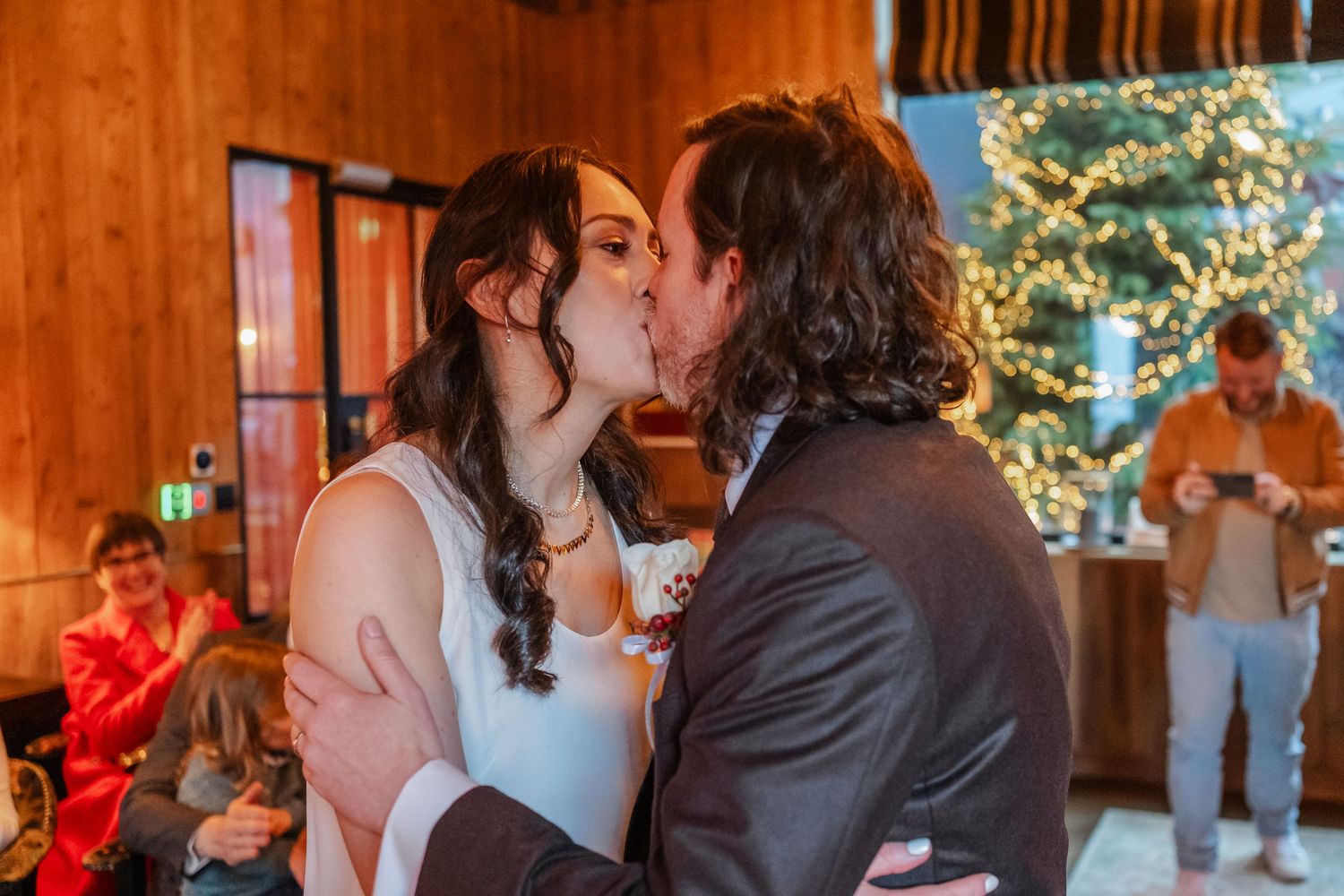 Couple sharing first kiss at wedding reception near Christmas tree in rustic wood-paneled venue.