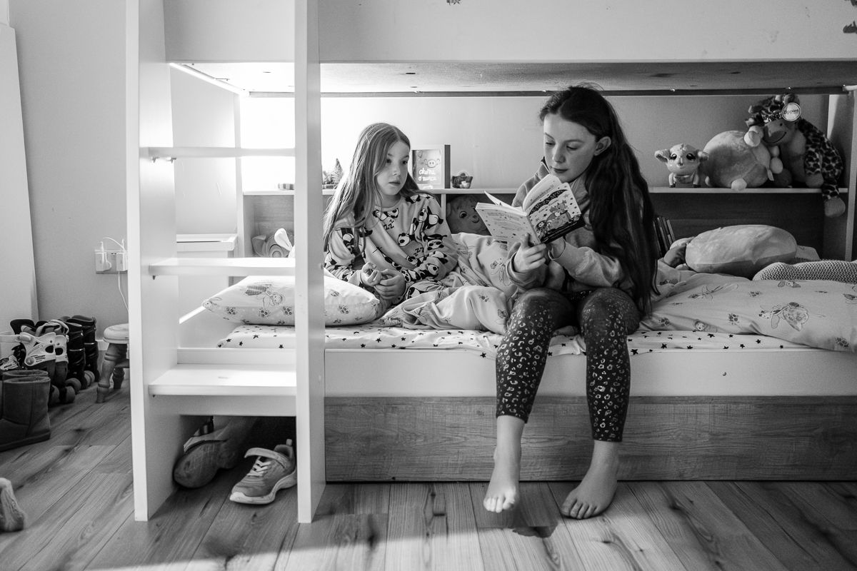 Two girls in pajamas sit on a bottom bunk bed reading a book together in a cozy bedroom.