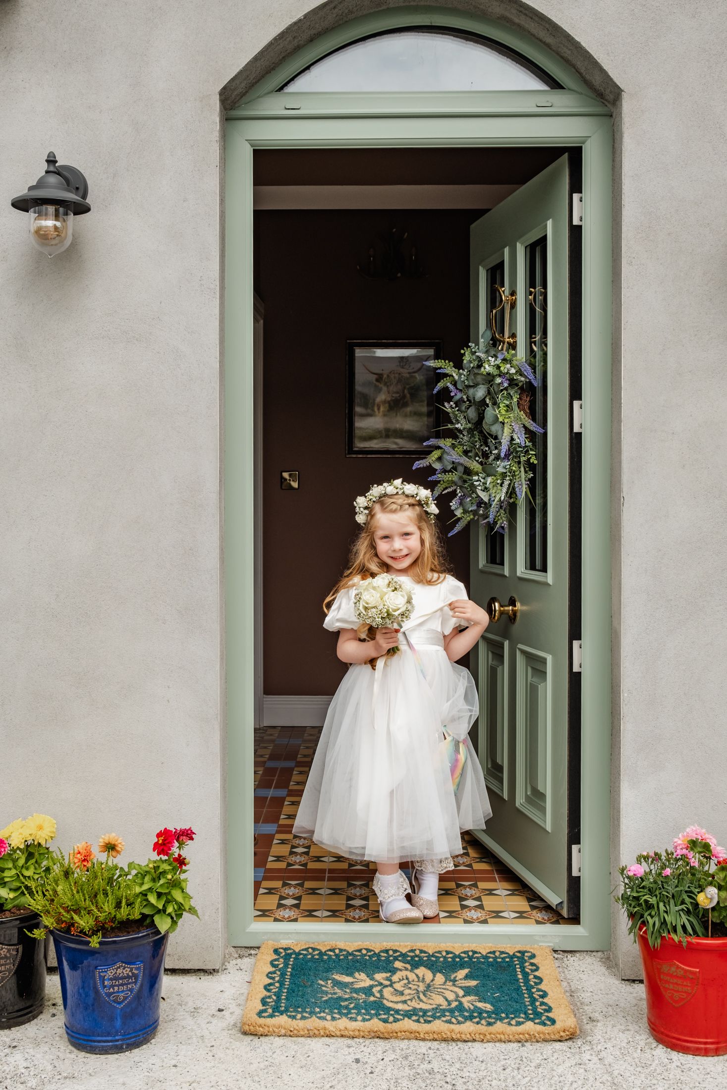 Small child in white dress and floral crown standing in elegant green doorway with potted flowers.