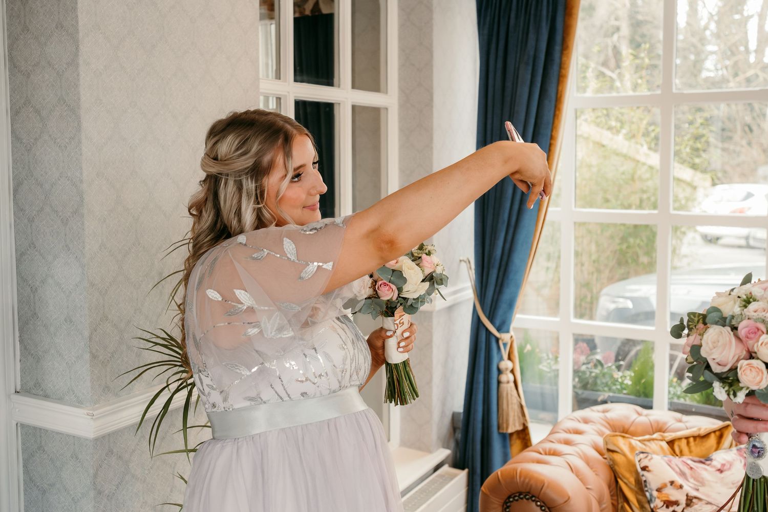Someone in a flowing dress reaches up to adjust curtains while holding a floral bouquet.