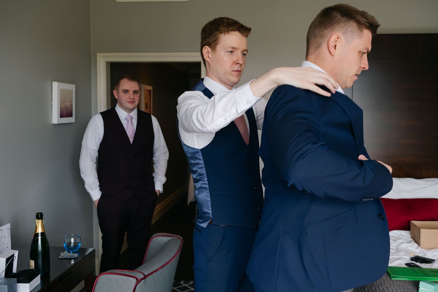 Three people in formal attire getting dressed in a room with a view of wine bottles and furnishings.