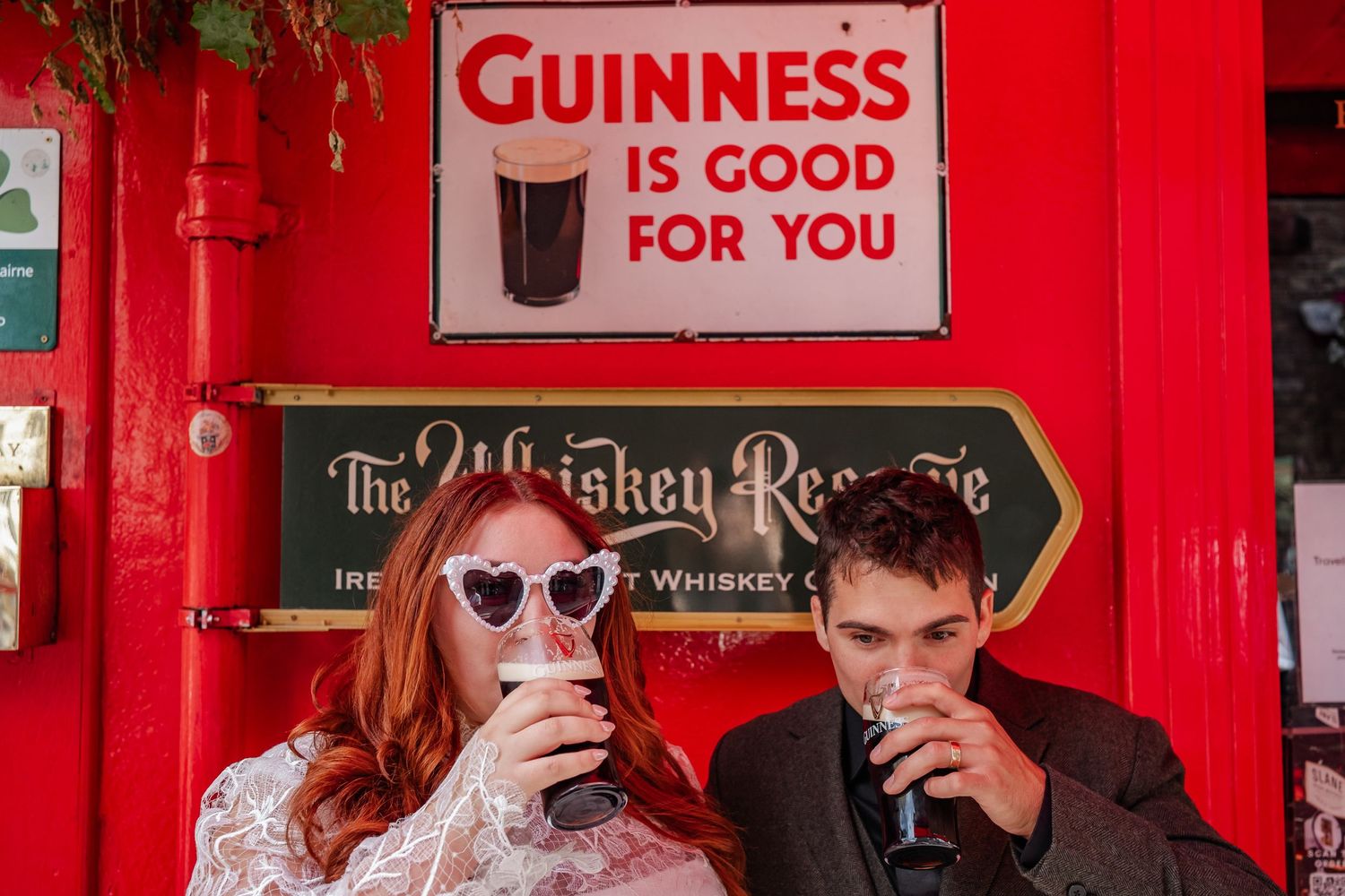 Couple drinking Guinness in front of vintage red pub wall with retro advertising sign.