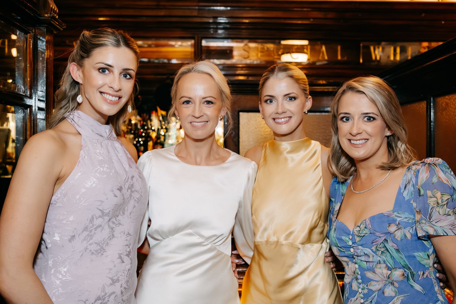 Four elegantly dressed women smiling together at a formal indoor event, wearing pastel gowns and floral attire.
