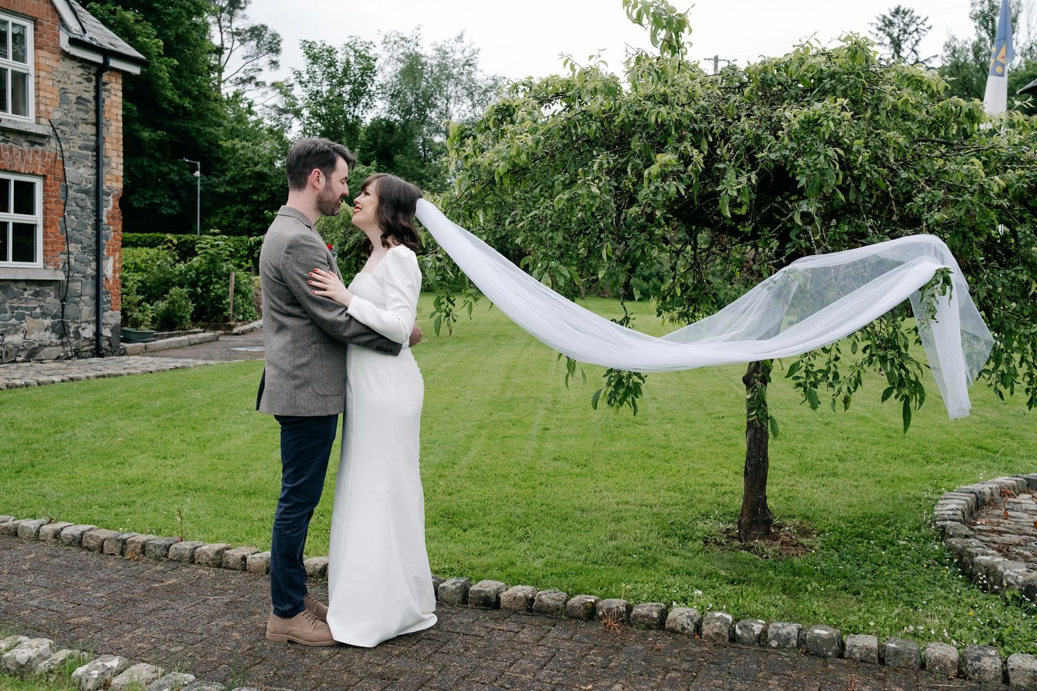 Bride and groom embrace outdoors with veil flowing in wind, lush green garden and stone building backdrop.