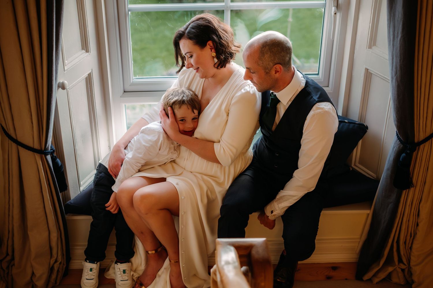 Bride and groom sit by a window with a child between them, sharing a tender family moment on their wedding day.