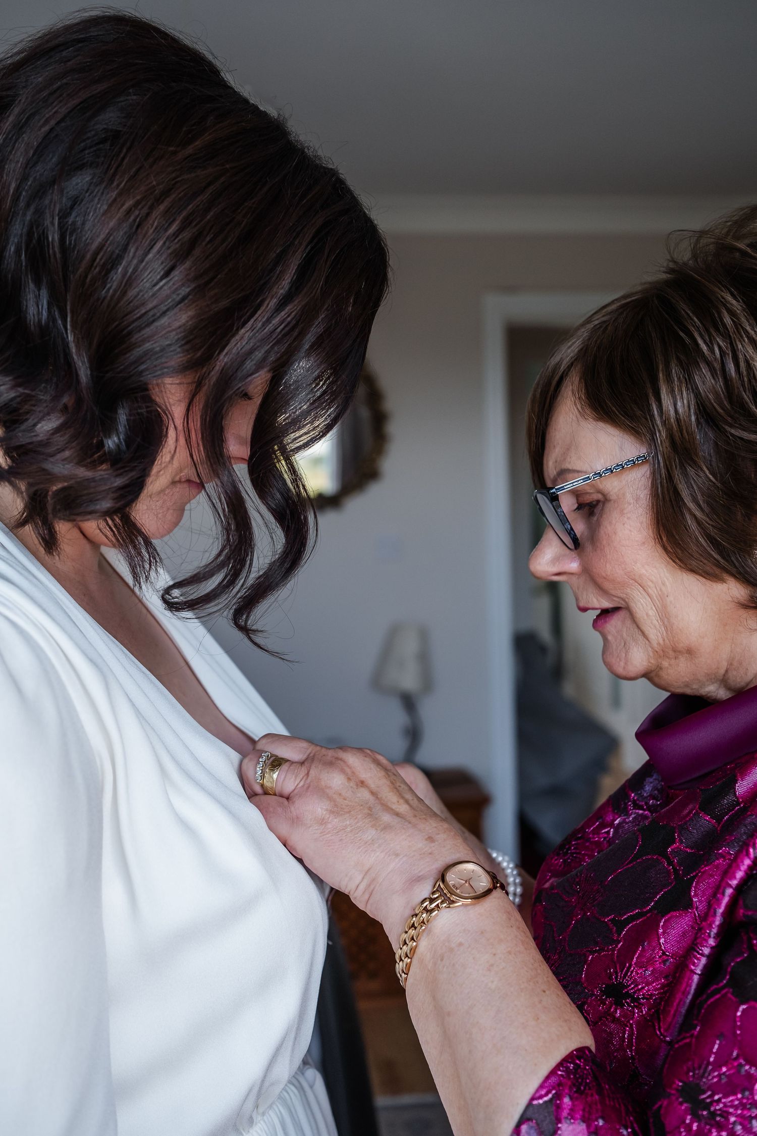 Mother helping bride with pearl necklace during wedding preparations, both wearing elegant attire.