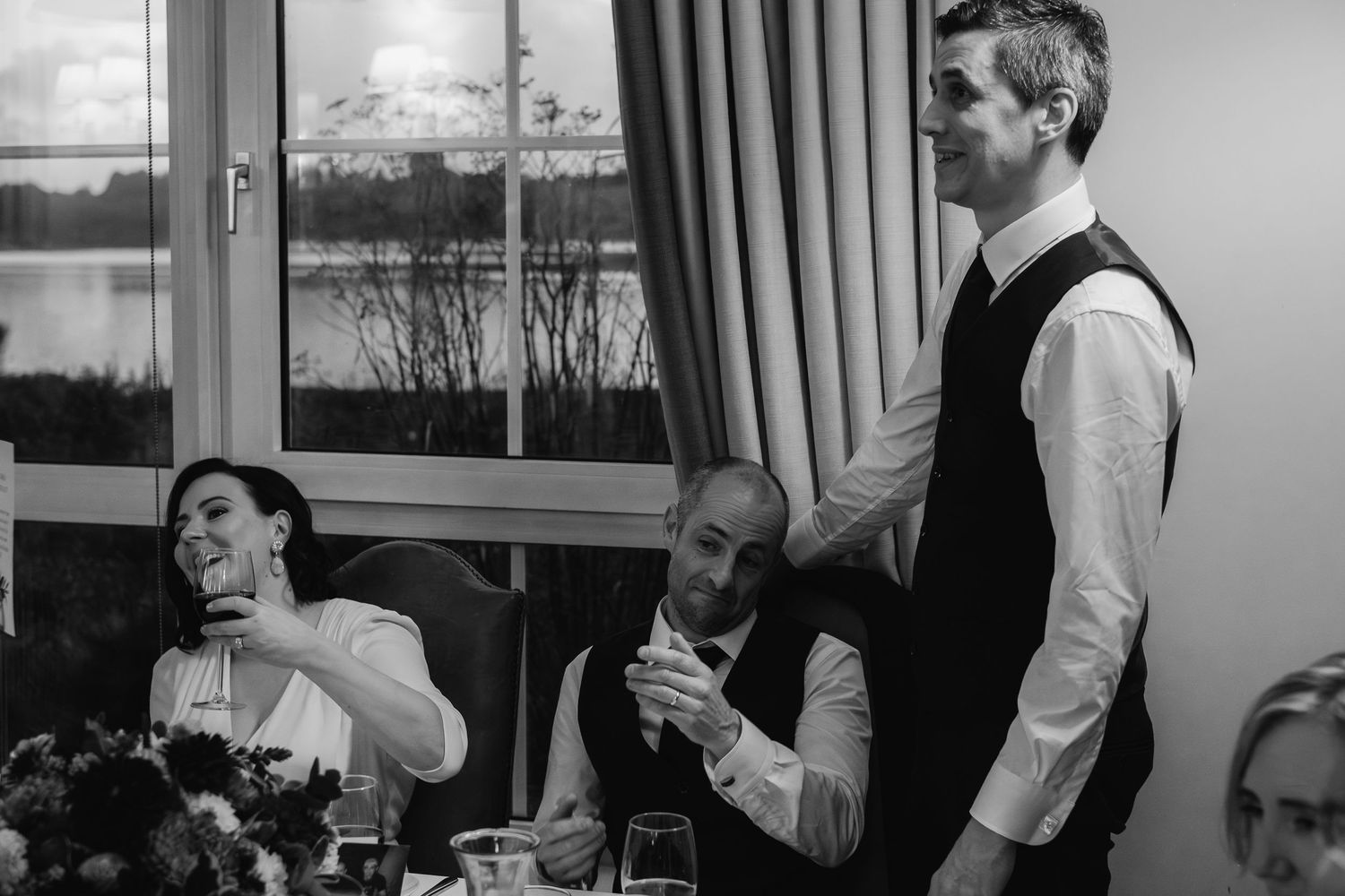Groom delivering a wedding speech while guests laugh at a reception dinner table.