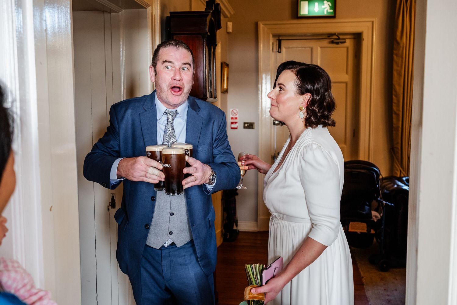 Surprised groom in blue suit holding a pint of beer with bride in white dress at wedding venue.