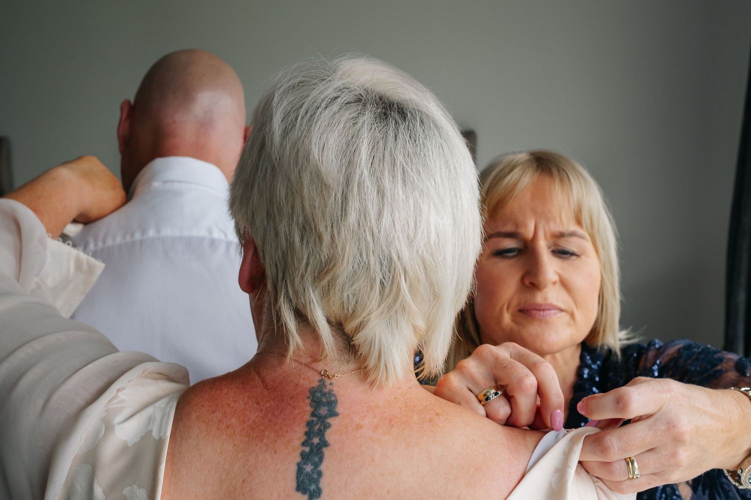 Wedding helper fastening bride's dress while groom stands nearby in white shirt.
