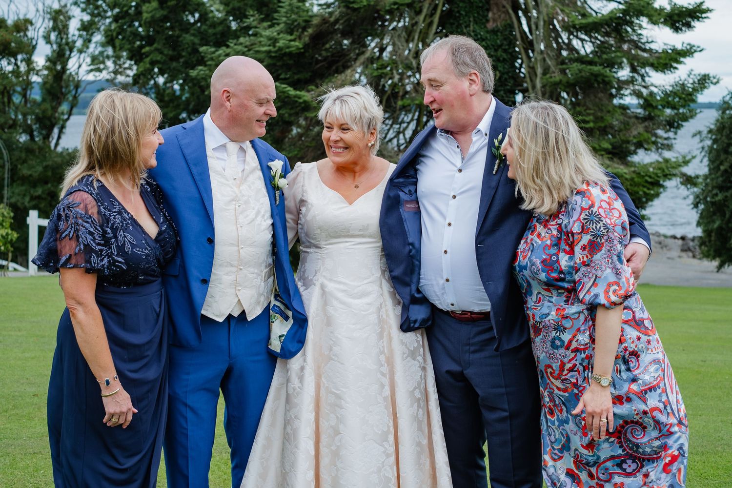 Wedding group photo with bride in white gown and groom in blue suit smiling with three guests outdoors.