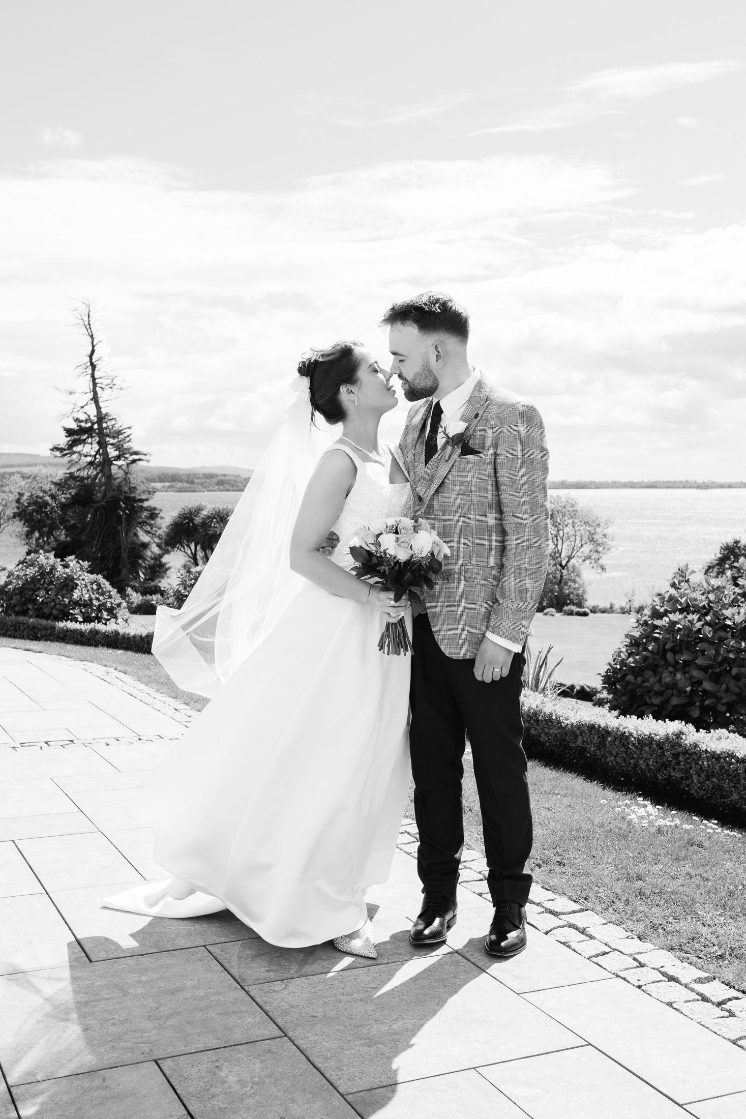 Black and white photo of a bride and groom sharing a romantic moment outdoors with scenic water views.