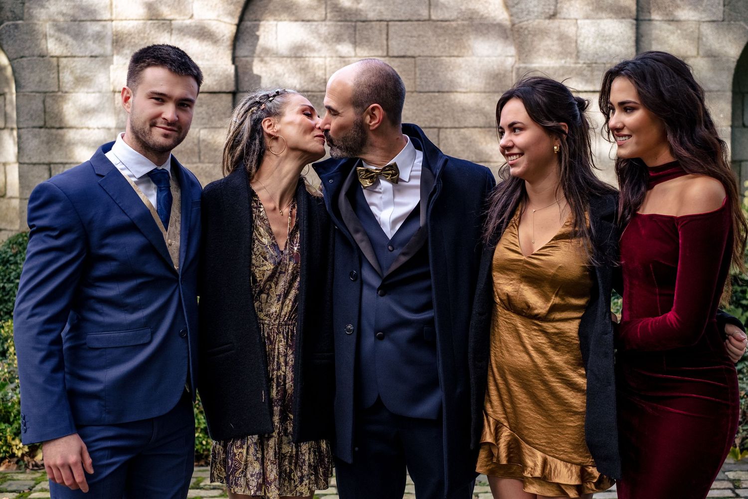 Five elegantly dressed people posing together at a wedding, with a couple sharing a kiss in the center.