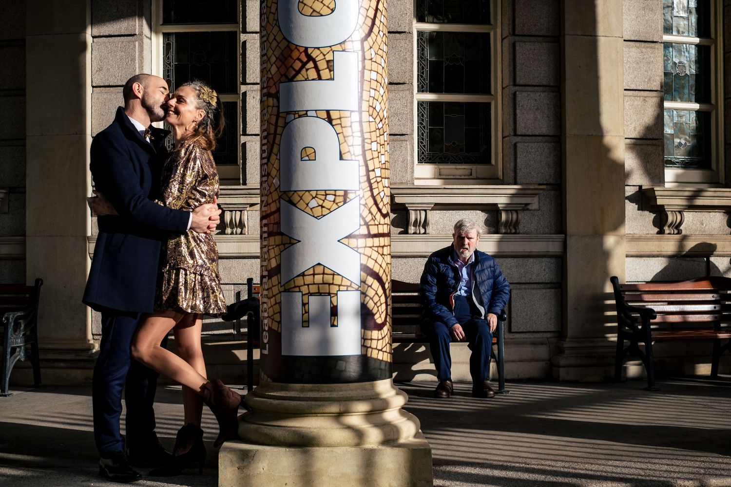A couple embraces near a decorative column outside a historic building, while an elderly man sits on a bench nearby.