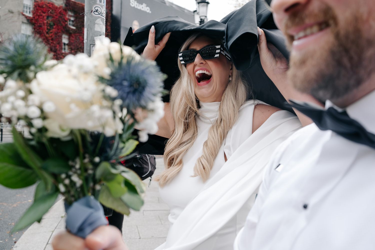 Laughing bride in white dress and black bow sunhat holds bouquet with groom in tuxedo on sunny wedding day.
