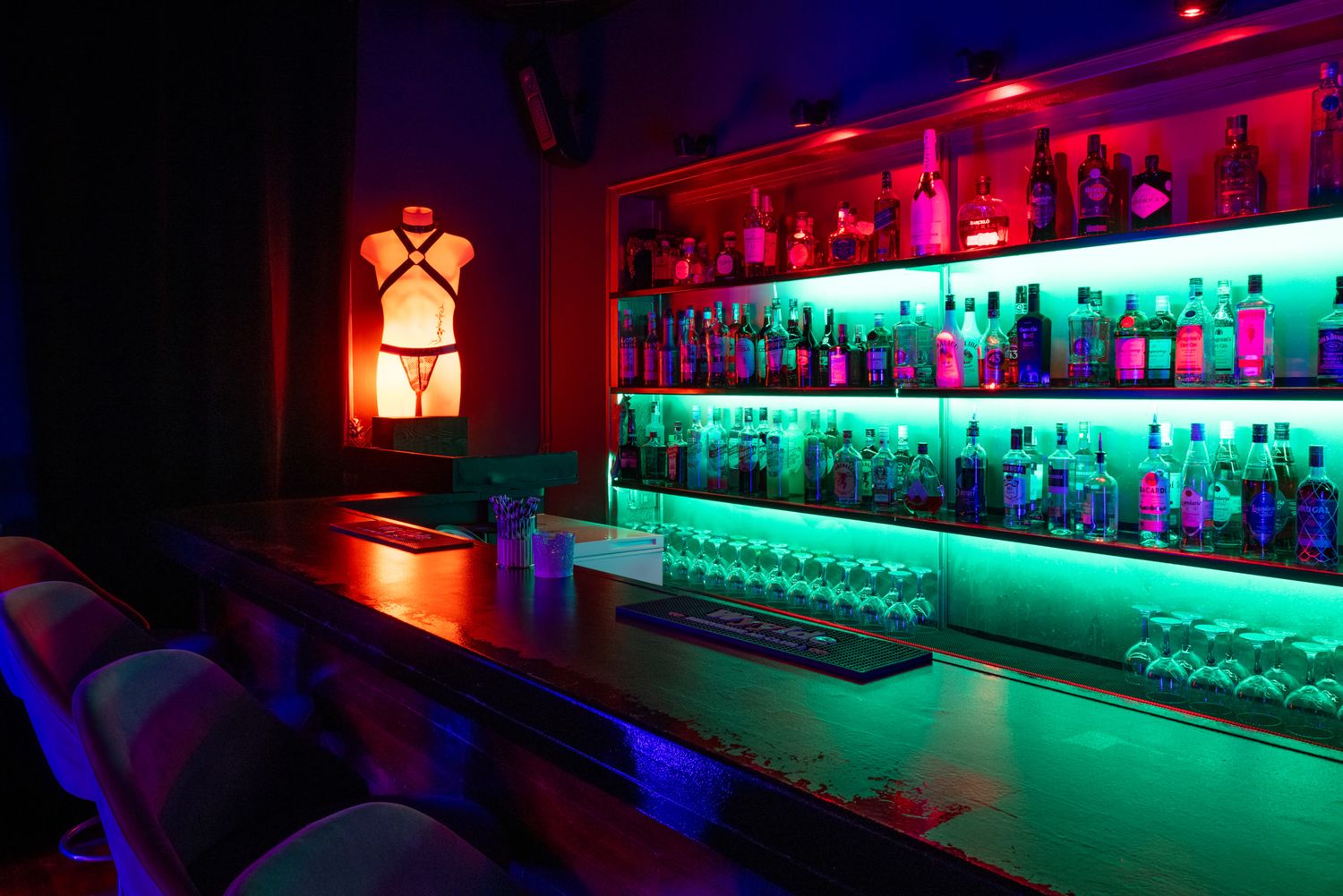 Dimly lit bar interior with red and green neon lighting illuminating rows of bottles on glass shelves behind the counter.