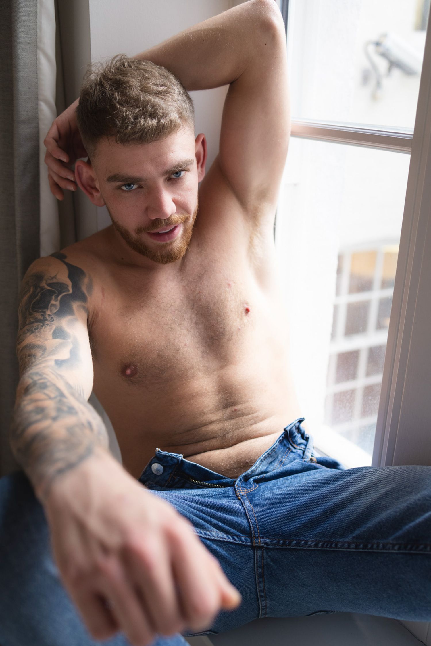 A person in blue jeans sits casually by a window showing a tattooed arm and posing for a dudeoir photoshoot.
