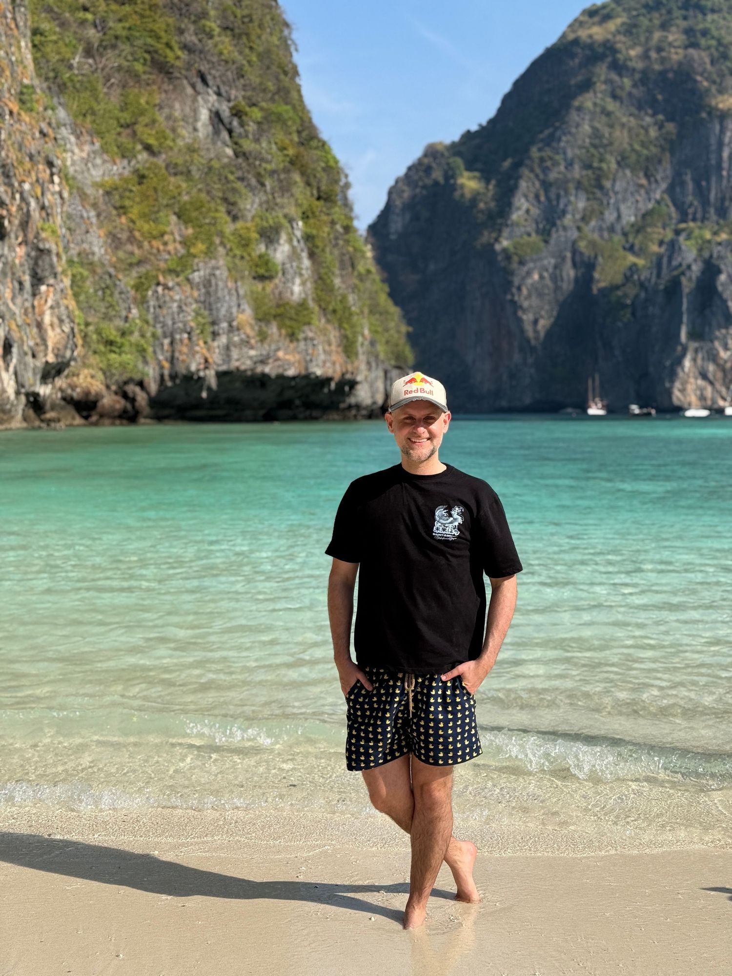 Man in black t-shirt and patterned shorts standing on tropical beach with turquoise water and limestone cliffs.