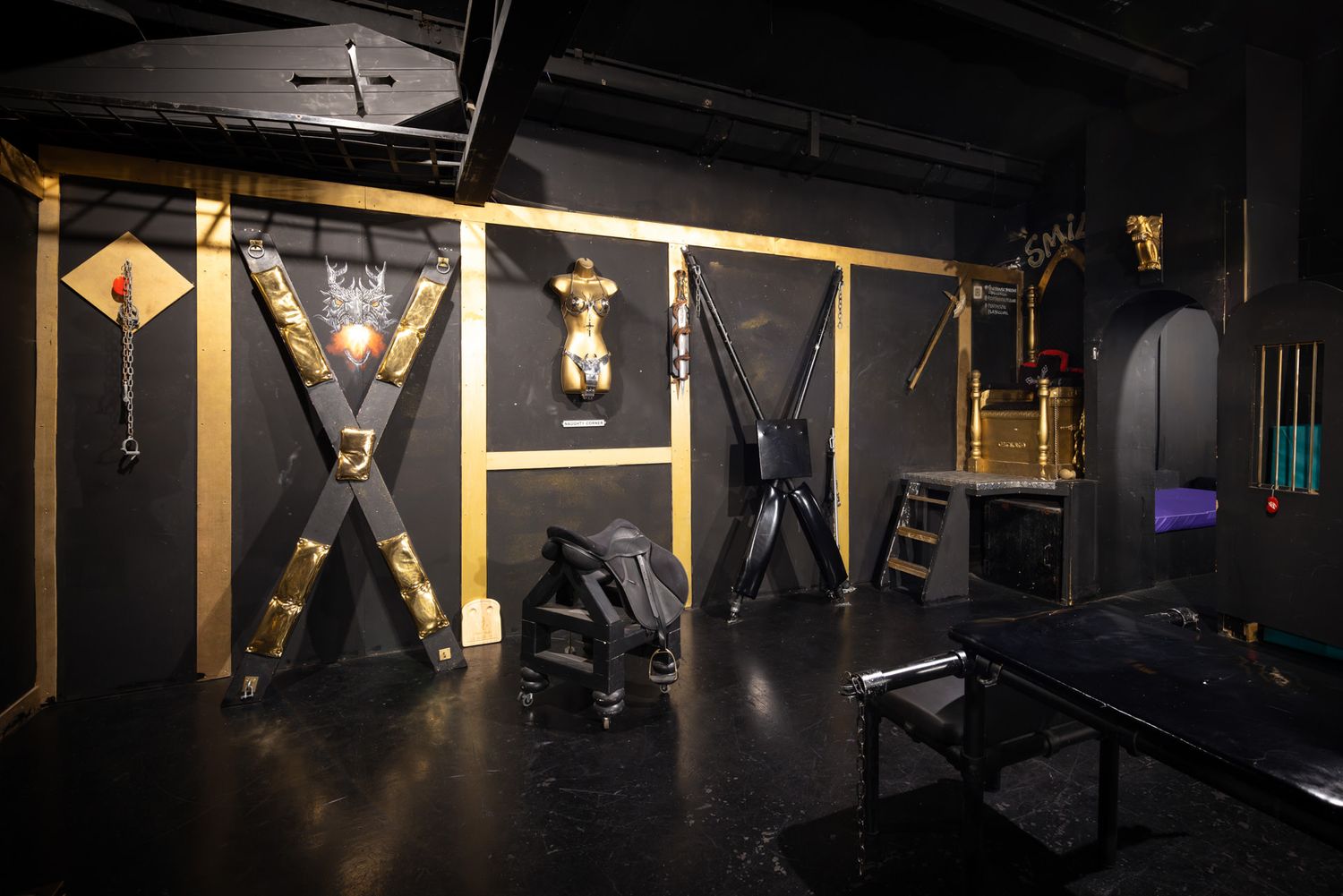 A dark BDSM dungeon room with wooden X-frame crosses, bondage equipment, and various restraints mounted on black walls.