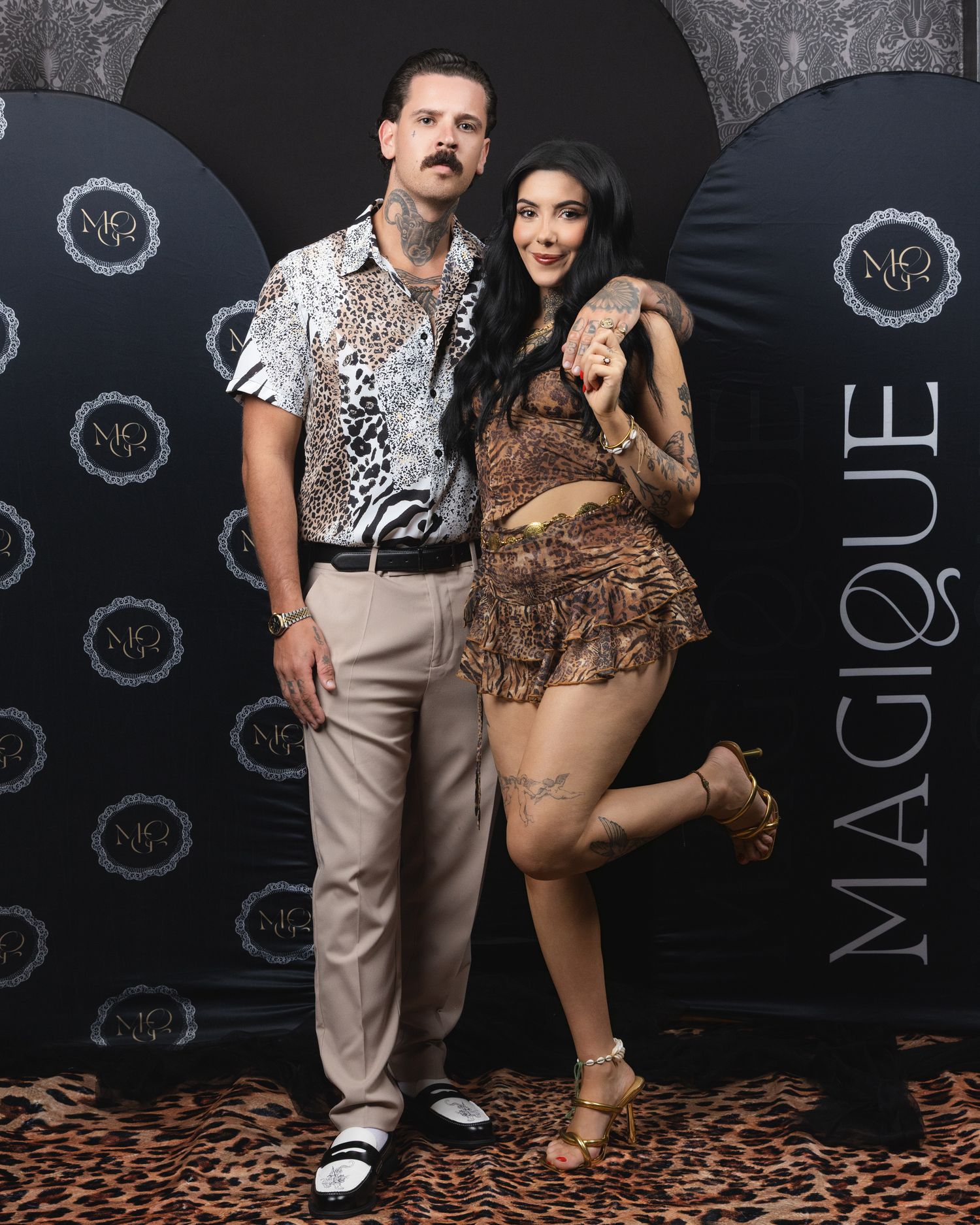 A tattooed couple poses at Magique event, man in leopard shirt and woman in brown animal print mini skirt outfit.